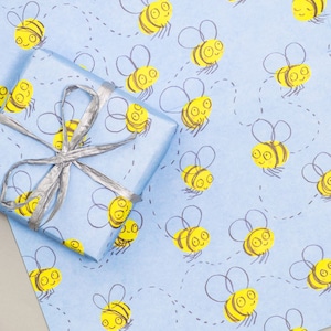 May include: Blue wrapping paper with a repeating pattern of yellow bees. The bees are drawn in a simple, cartoon style and are flying in different directions. A silver ribbon is tied around a gift box on top of the wrapping paper.