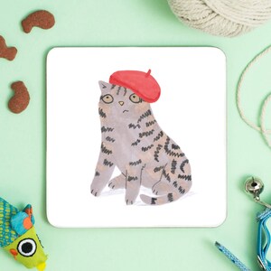 May include: A gray and brown tabby cat wearing a red beret. The cat is sitting on a white background.