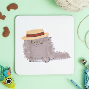 May include: A gray cat with a fluffy coat wears a straw hat with a red and black band. The cat has a grumpy expression on its face.