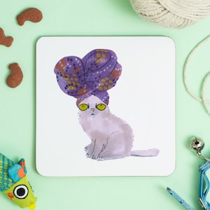 Könnte beinhalten: Eine weiße Katze mit gelben Augen trägt einen turban mit violettem und goldenem Muster. Die Katze sitzt auf einem weißen Hintergrund.