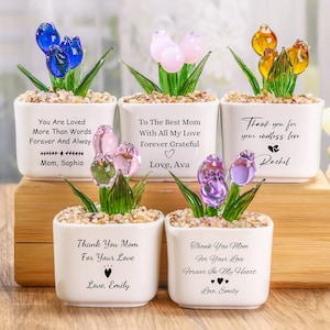 Puede incluir: Una colección de pequeños maceteros de cerámica blanca, cada uno con un arreglo de tulipanes de cristal. Los tulipanes son de varios colores: azul, rosa, amarillo y morado. Cada maceta tiene un mensaje diferente, como "You Are Loved" o "Thank You Mom".