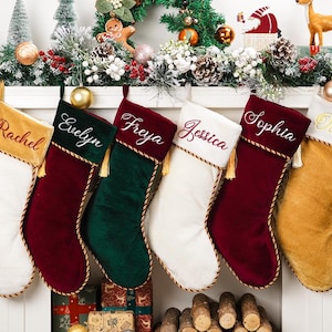 Puede incluir: Seis calcetines navideños personalizados en varios colores, incluyendo blanco, rojo, verde y dorado. Cada calcetín tiene un nombre bordado en un color complementario, con borla y ribete dorados. Decoración festiva navideña en el fondo.
