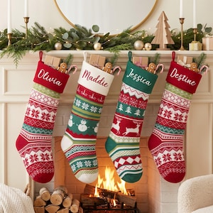 May include: Four personalised Christmas stockings hanging in a row. Two are red with white cuffs and two are white or green with contrasting cuffs. Each features knit patterns including snowflakes, reindeer, and snowmen, with names Olivia, Maddie, Jessica, and Destine.