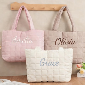 May include: Three quilted tote bags in light pink, beige, and white. Each bag features a rectangular grid pattern, two shoulder straps, and a name embroidered in cursive script on the front: Amelia in white, Olivia in brown, and Grace in blue.