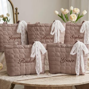 Puede incluir: Cinco bolsos tote acolchados con estampado de cuadros vichy en marrón y blanco, cada uno con un nombre bordado en hilo marrón oscuro y un lazo de encaje blanco atado a un asa. Los nombres son Harper, Meuve, Sophia, Rachel y Grace.
