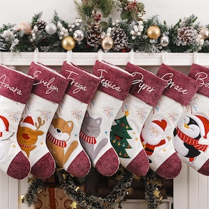 Puede incluir: Siete calcetines navideños blancos con puños de color burdeos texturizado, cada uno personalizado con un nombre en letra blanca. Los calcetines presentan coloridos gráficos navideños bordados que incluyen un muñeco de nieve, reno, zorro, gato, árbol, Santa y pingüino.