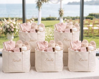 May include: Five off-white canvas tote bags, each embroidered with a name: Jessie, Olivia, Sarah, Emily, or Laura. Each bag contains a folded dusty pink robe, a pair of beige open-toe slippers, a small pink jewelry box, and a white bottle.
