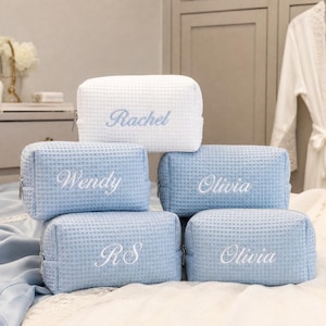 Puede incluir: Una colección de cinco neceseres rectangulares con textura de gofre y cierres de cremallera. Uno es blanco con bordado azul que dice Rachel, mientras que cuatro son azul claro con bordado blanco que dice Wendy, Olivia, RS y Olivia.