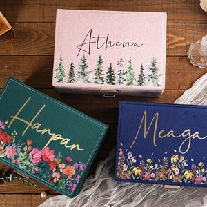May include: Three personalized jewelry boxes in pink, teal, and navy blue. The pink box has the name "Athena" and a forest design. The teal box has the name "Harpan" and a floral design. The navy box has the name "Meagan" and a floral design.