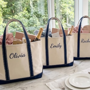 Op de afbeelding: Drie off-white canvas draagtassen met marineblauwe randen en hengsels. Elke tas is voorzien van een in marineblauw cursief geborduurde naam: Olivia, Emily en Rachel. De tassen zijn gevuld met diverse geschenken, waaronder flessen.
