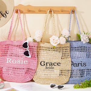 Könnte beinhalten: Drei offene Tragetaschen in Rosa, Beige und Blau, jede mit geflochtenem Henkel. Die rosa Tasche trägt die Aufschrift "Rosie", die beige Tasche "Grace Bride" und die blaue Tasche "Paris". Jede Tasche hat Blumen.