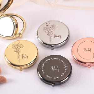 Könnte beinhalten: Eine Sammlung personalisierter Kompaktspiegel in Gold, Silber und Roségold. Jeder runde Spiegel hat ein Blumendesign und einen Namen. Ein Spiegel ist geöffnet und zeigt eine reflektierende Oberfläche. Make-up-Pinsel sind sichtbar.