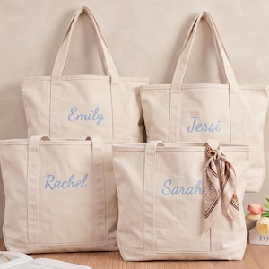 May include: Four off-white canvas tote bags with two shoulder straps each. Each bag features a name embroidered in light blue script: Emily, Jessi, Rachel, and Sarah. The bag labeled Sarah includes a patterned brown and white silk-style scarf tied to one handle.