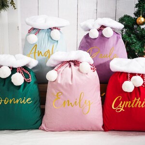 Könnte beinhalten: Fünf farbenfrohe Weihnachtsgeschenktüten in Grün, Rosa, Rot, Lila und Blau. Jede Tasche hat einen weißen, flauschigen Besatz, weiße Pompons und einen goldenen Namen. Die Taschen sind mit rot-weißem Seil gebunden. Im Hintergrund ist ein Weihnachtsbaum.