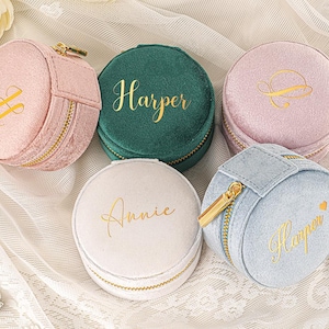Könnte beinhalten: Eine Sammlung runder Schmuckkästchen aus Samt in verschiedenen Pastellfarben: Rosa, Grün, Hellrosa, Weiß und Blau. Jedes Etui hat einen goldenen Reißverschluss und personalisierte goldene Schrift. Auf einer weißen Spitzentischdecke angeordnet.