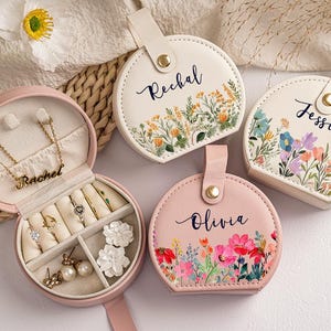 May include: A pink, round jewelry travel case with a gold necklace and rings inside. Three additional round cases with floral designs and names "Rechal", "Jessica", and "Olivia" are also shown. The cases have a button closure and a small handle.