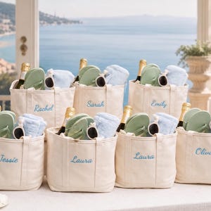 Puede incluir: Siete bolsos de lona color hueso, cada uno bordado con un nombre único en letra azul. Cada bolso contiene una botella de champán, un par de chanclas verde claro, gafas de sol con montura blanca y una toalla doblada de color azul claro.