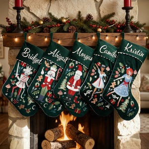 Op de afbeelding: Vijf donkergroene kerstsokken hangen op een rij, elk gepersonaliseerd met wit geborduurde namen: Bridget, Abigail, Morgan, Camila en Kristin. De sokken zijn voorzien van feestelijke kerstillustraties zoals een notenkraker, de Kerstman en dansers, geaccentueerd met peperkoekmannetjes, zuurstokken en dennentakjes.