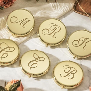 May include: Six round, gold-toned metal compact mirrors, each featuring a single engraved script letter on the lid. The letters M, R, K, C, E, and B are displayed in a decorative, flowing font across the polished, reflective surfaces.