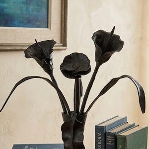 May include: A wrought iron sculpture of calla lilies in a vase. The black metal flowers are displayed on a wooden table with books and a compass. The books include titles like "Wrought Iron Artistry" and "Botanical Forms."