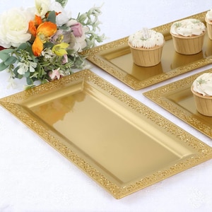 May include: Gold rectangular serving trays with ornate, embossed borders. The trays are empty, and some are holding cupcakes with white frosting. A bouquet of flowers is in the background. These trays are suitable for serving food at a party or event.