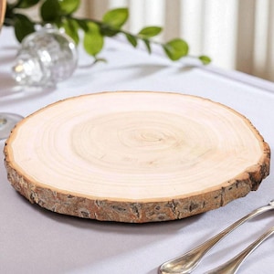 May include: A round, natural wood slice with a rough bark edge. The light-colored wood grain is visible across the surface. The wood slice is on a white tablecloth, with silverware and a champagne glass in the background.