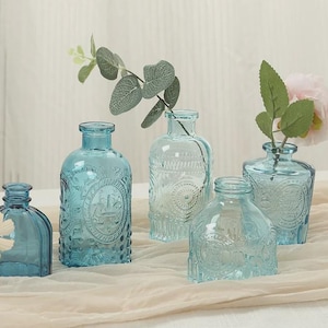 May include: A collection of five small blue glass bottles with embossed patterns. Each bottle has a narrow neck and a distinct shape. Some bottles contain small artificial flowers or green leafy stems, creating a delicate display of blue glass.