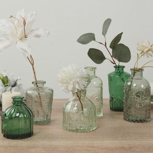 May include: A collection of five small glass vases in varying shades of green, featuring embossed patterns and textured surfaces. Some vases contain artificial flowers, including a white lily, a white chrysanthemum, and eucalyptus stems.