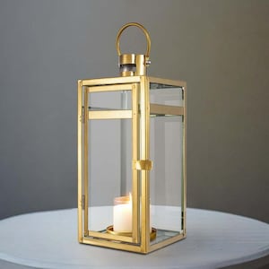 May include: A gold-colored metal lantern with clear glass panels and a handle. A lit white candle sits inside the lantern, casting a warm glow. The lantern is rectangular and sits on a white table.