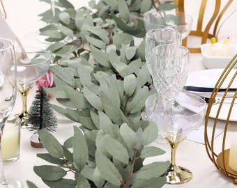 May include: A table setting featuring a eucalyptus garland centerpiece. Clear stemmed glasses with gold accents are arranged around the garland. A small Christmas tree and candle are also visible, creating an elegant and festive atmosphere.