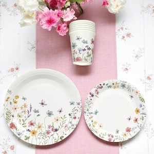 May include: A set of floral-themed disposable tableware, including two plates and a stack of cups. The plates and cups feature a delicate design of colorful wildflowers and butterflies. The background includes a pink cloth and a floral patterned surface.