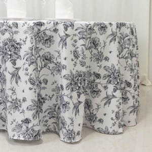 May include: A round white tablecloth with a black floral pattern. The design features detailed botanical illustrations of flowers and foliage, creating a classic and elegant aesthetic. The tablecloth is suitable for a dining table.