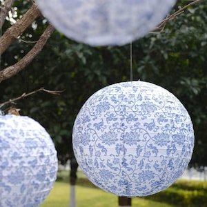 May include: Three round paper lanterns with a blue and white floral pattern hang from tree branches. The lanterns are suspended by thin wires, with a blurred green background of foliage.