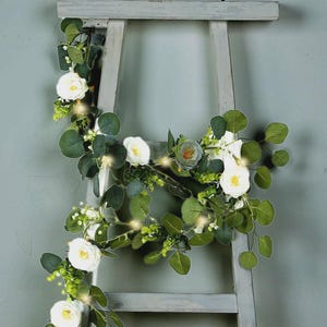 May include: A decorative garland featuring white roses, green eucalyptus leaves, and small white flowers. The garland is draped over a gray wooden ladder and includes small, warm-toned lights, creating a soft, inviting ambiance. Perfect for home decor.