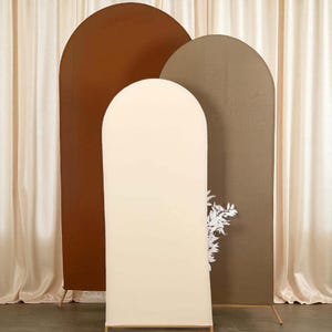 May include: Three arched backdrop panels in shades of brown and cream. The panels are made of fabric and are set on gold-colored metal frames. A white floral arrangement is partially visible on one panel. The backdrop is set against a cream-colored curtain.