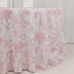 May include: A white tablecloth with a pink floral pattern. The design features detailed illustrations of flowers and foliage, creating a classic and elegant aesthetic. The tablecloth is draped over a round table, showcasing the fabric's texture and design.