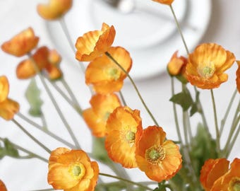 May include: A close-up of a bouquet of orange poppy flowers with green stems and leaves. The flowers have a yellow center and are arranged in a vase. A white plate and fork are in the background.