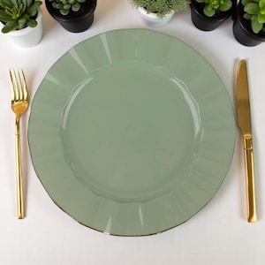 May include: A sage green ceramic plate with a ruffled edge, paired with gold-colored fork and knife. The plate has a subtle gold trim. The arrangement is set on a white surface, with small potted plants in the background.