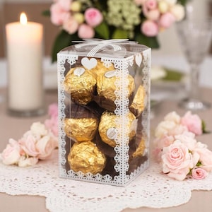 May include: A clear, square gift box filled with gold-wrapped chocolates. The box has a white lace-like design and a white ribbon. The chocolates are arranged inside the box, and the background includes a lit candle and pink roses.