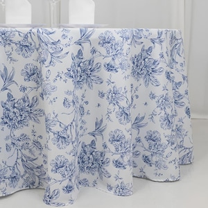 May include: A white tablecloth with a blue floral pattern. The design features detailed illustrations of flowers and foliage, creating a classic and elegant aesthetic. The tablecloth is draped over a round table, with plates and napkins visible.