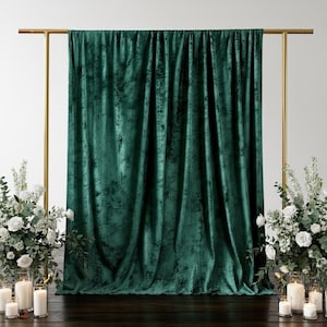 May include: A dark emerald green velvet curtain hangs from a gold metal frame. The fabric is draped, creating soft folds and a textured appearance. White floral arrangements and candles flank the curtain, creating a decorative backdrop.