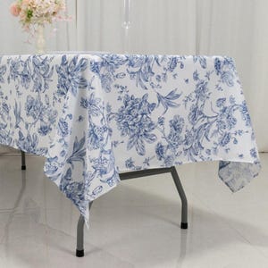May include: A white tablecloth with a blue floral pattern covers a table. The floral design features detailed illustrations of flowers and foliage. The tablecloth is draped over a metal folding table, creating a decorative and functional surface for dining or events.