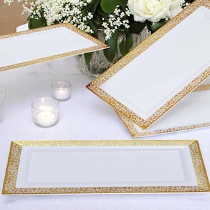 May include: White rectangular serving platters with gold decorative borders. The platters are arranged on a white tablecloth, with candles and a floral arrangement in the background. The gold border has a repeating floral design.
