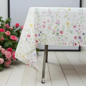 May include: A white tablecloth with a delicate floral pattern featuring pink, yellow, and purple wildflowers. The tablecloth is draped over a table with a silver leg, creating a fresh and inviting look. The floral design adds a touch of spring to any dining setting.