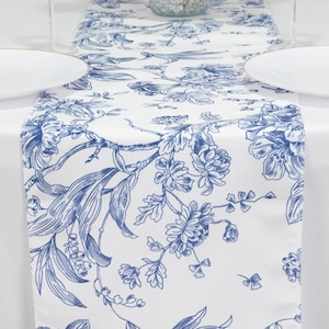 May include: White table runner with a blue floral design. The runner features a repeating pattern of flowers, leaves, and branches. The design is in a classic blue and white color scheme, perfect for adding a touch of elegance to any dining table.