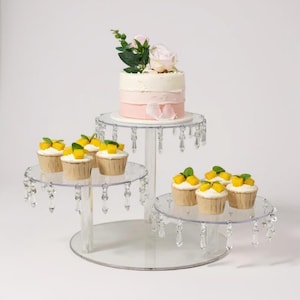 May include: A clear, three-tiered cake stand with hanging crystal accents. The top tier holds a small cake with pink and white frosting and a floral arrangement. Cupcakes with yellow fruit toppings are on the lower tiers.
