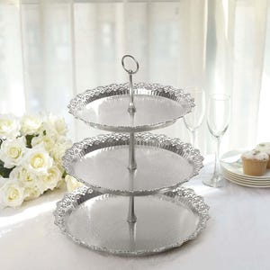 May include: A three-tiered silver serving tray with ornate, scalloped edges. The tray has a central handle and is designed for displaying desserts or appetizers. The background includes white flowers and champagne glasses.