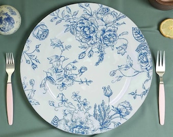 May include: White ceramic plate with a blue floral design. The plate features a detailed illustration of roses and other flowers. Two forks with pink handles are on either side of the plate. The plate is on a green surface.
