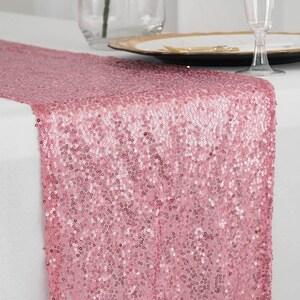 May include: A pink sequin table runner. The runner is made of small, shiny sequins that catch the light. It is draped over a white tablecloth, with a plate and glass in the background. The runner adds a touch of glamour to any table setting.