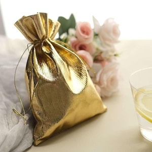 May include: A shiny gold drawstring gift bag with a textured surface. The bag is tied with a gold string and sits next to a bouquet of pink roses and a glass of water with lemon slices.